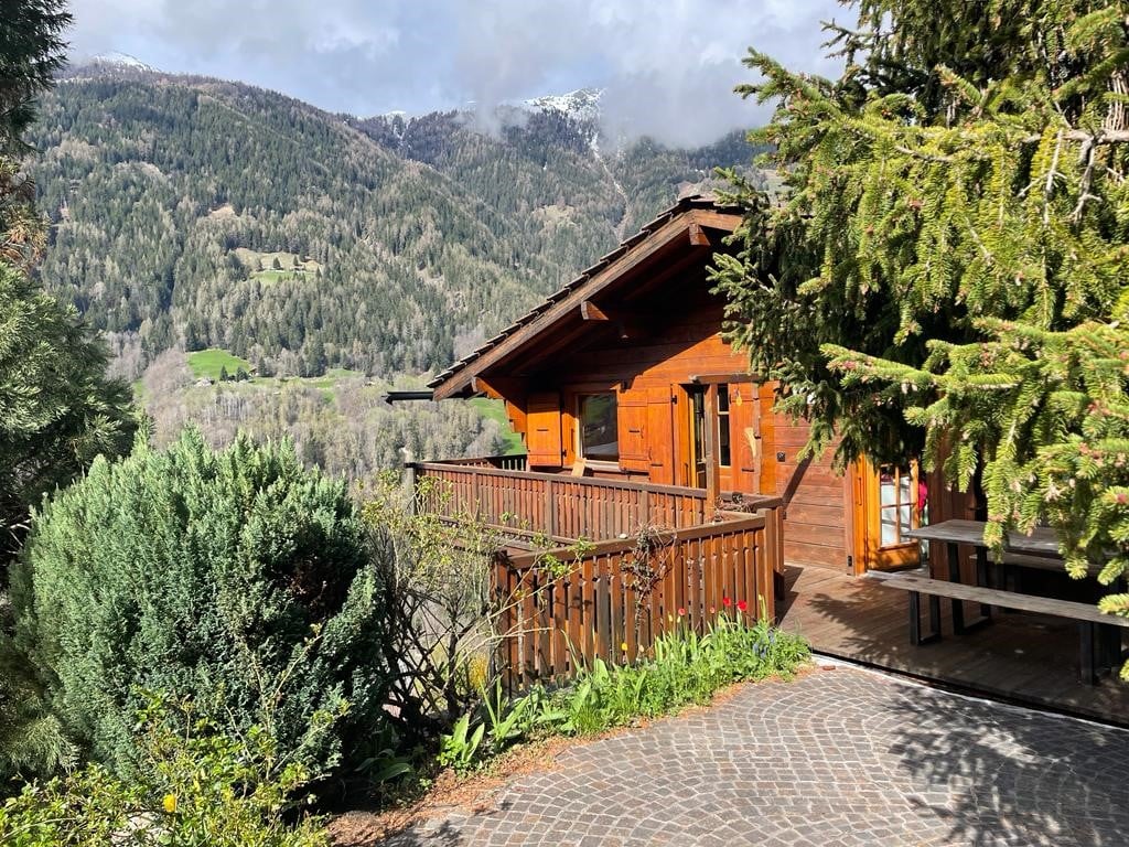 A traditional wooden chalet is framed by lush greenery and flowering plants. A wrap-around balcony offers scenic views of the surrounding mountains and valley. The entryway features wooden doors that complement the rustic charm of the structure.