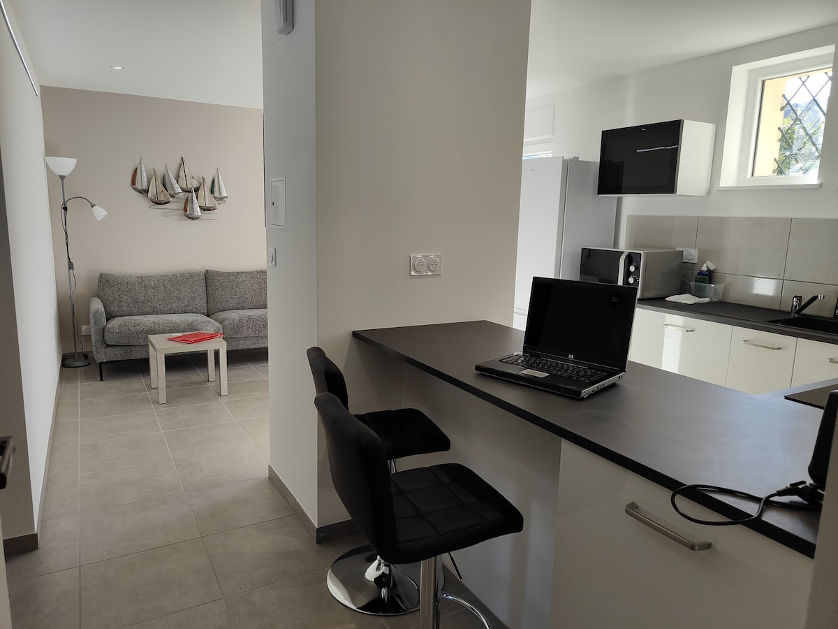 A modern open-concept space is presented, featuring a kitchen area with a bar countertop and two stools. A living area is visible, highlighted by a grey sofa and decorative wall art depicting sailboats. Natural light enters through a nearby window.