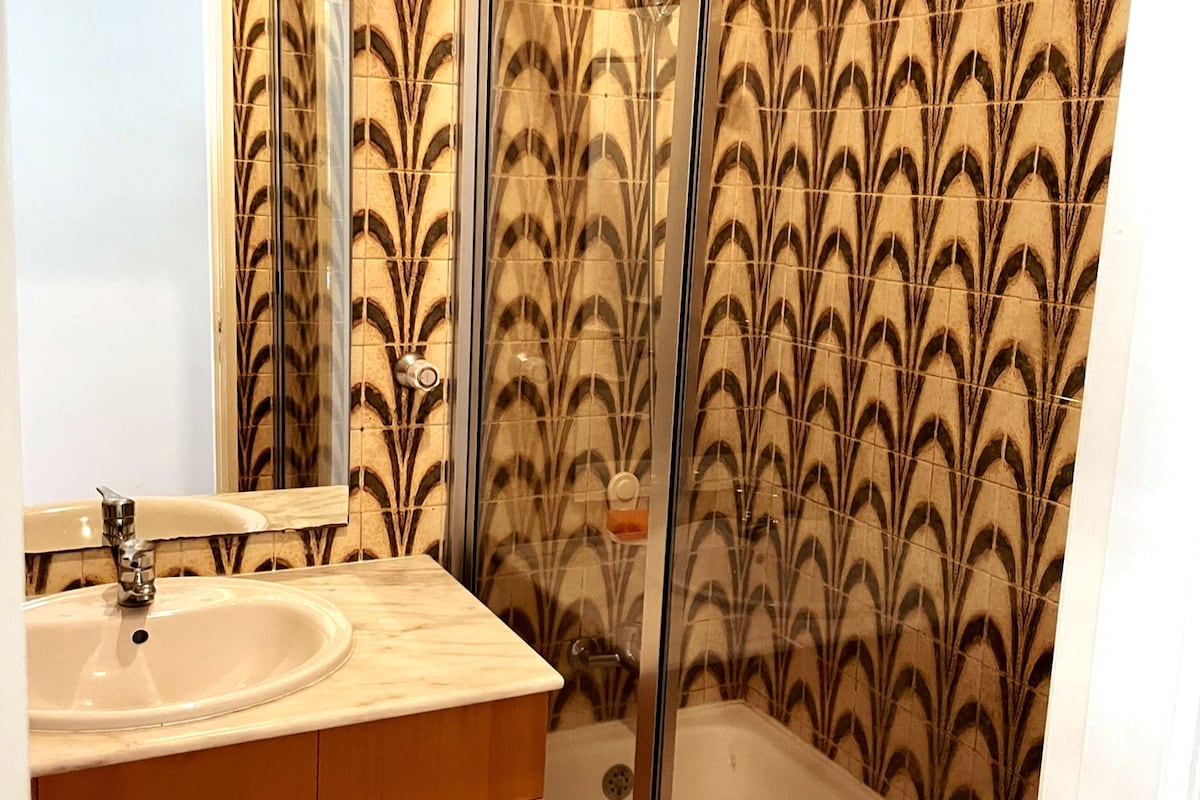 A shared bathroom features a shower enclosed in glass, surrounded by patterned tiles in warm tones. A sink with a marble countertop and a mirror are visible, providing a functional and stylish space for guests.