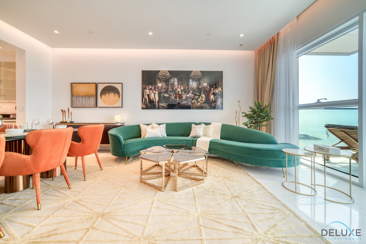 The living area is enhanced by a modern curved green sofa, complemented by stylish orange chairs. A soft area rug anchors the space, and a large artwork adorns the wall, depicting a gathering. Natural light floods in from large windows, revealing a view of the sea.
