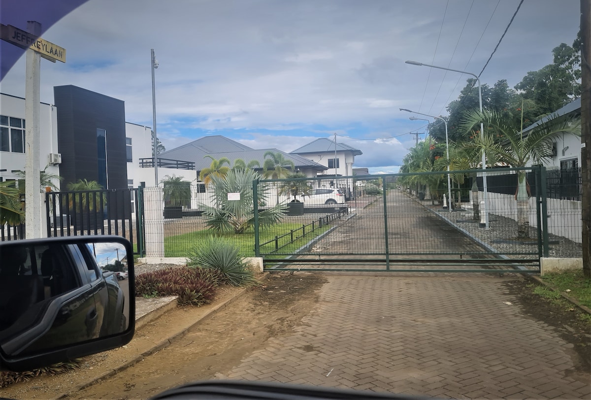 A gated entrance is shown, featuring a well-maintained driveway lined with palm trees. The modern buildings are visible in the background, set against a partly cloudy sky. A secure atmosphere is conveyed through the gate and surrounding fences.