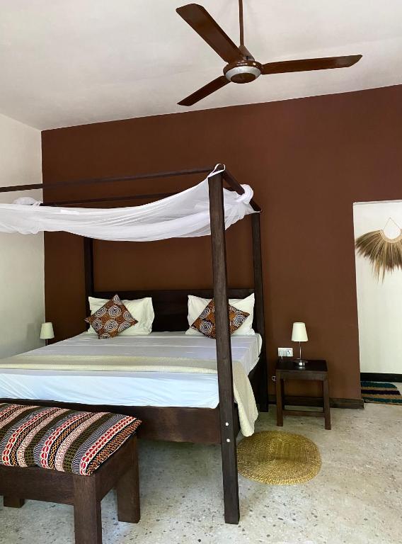 A tranquil bedroom features a dark wood four-poster bed draped with white fabric. Accent pillows are arranged on the bed, complemented by a patterned ottoman at its foot. A light fixture is positioned on each side, while a ceiling fan provides airflow.