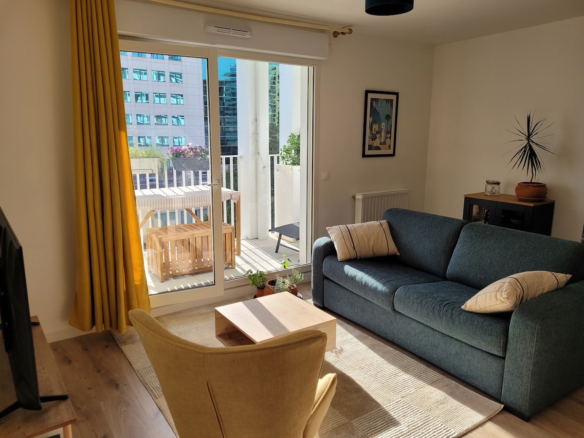 A bright living area features a comfortable sofa with decorative cushions facing a balcony door. Natural light floods the space, highlighting a small wooden table and a cozy armchair. A soft area rug lies beneath, creating a warm and inviting setting.