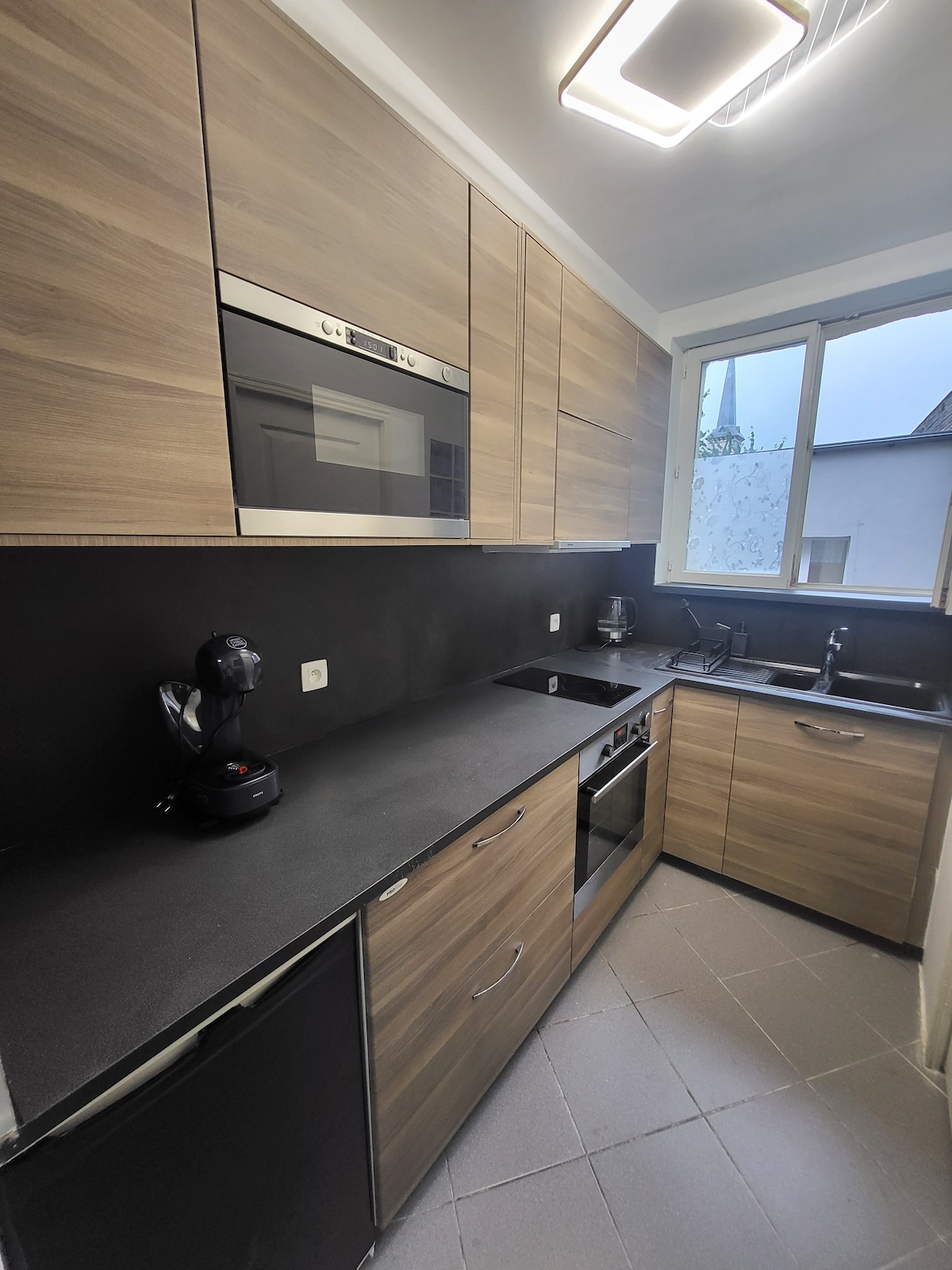 A modern kitchen is presented with sleek wooden cabinetry, featuring built-in appliances including an oven, stove, and microwave. A coffee machine is placed on the counter, and a window provides natural light, illuminating the contemporary design of the space.
