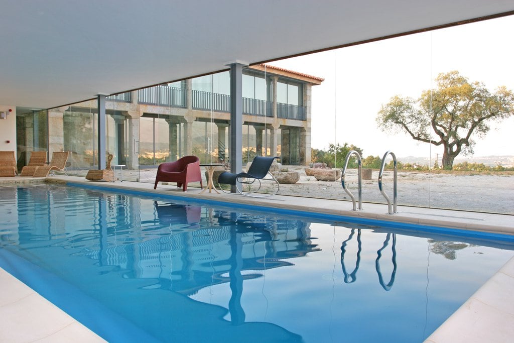 An indoor heated pool glistens under natural light, surrounded by large glass walls that provide an unobstructed view of the outdoors. Comfortable seating options, including a red armchair and a blue lounge chair, are positioned nearby, enhancing the serene environment.