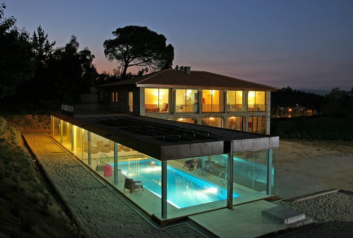 The two-story structure is illuminated at dusk, with large glass windows showcasing the inviting interiors. A spacious outdoor area features a heated swimming pool, surrounded by lounge seating and lush vegetation, seamlessly blending the tranquility of nature with modern design.