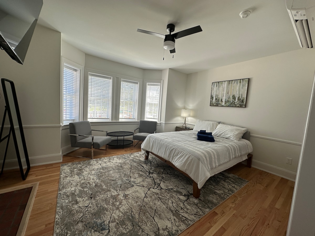 A bright and airy studio features a spacious bed with crisp linens, flanked by large bay windows that allow ample natural light. Two modern chairs and a small table create a comfortable seating area, while a soft area rug adds warmth to the wooden flooring.