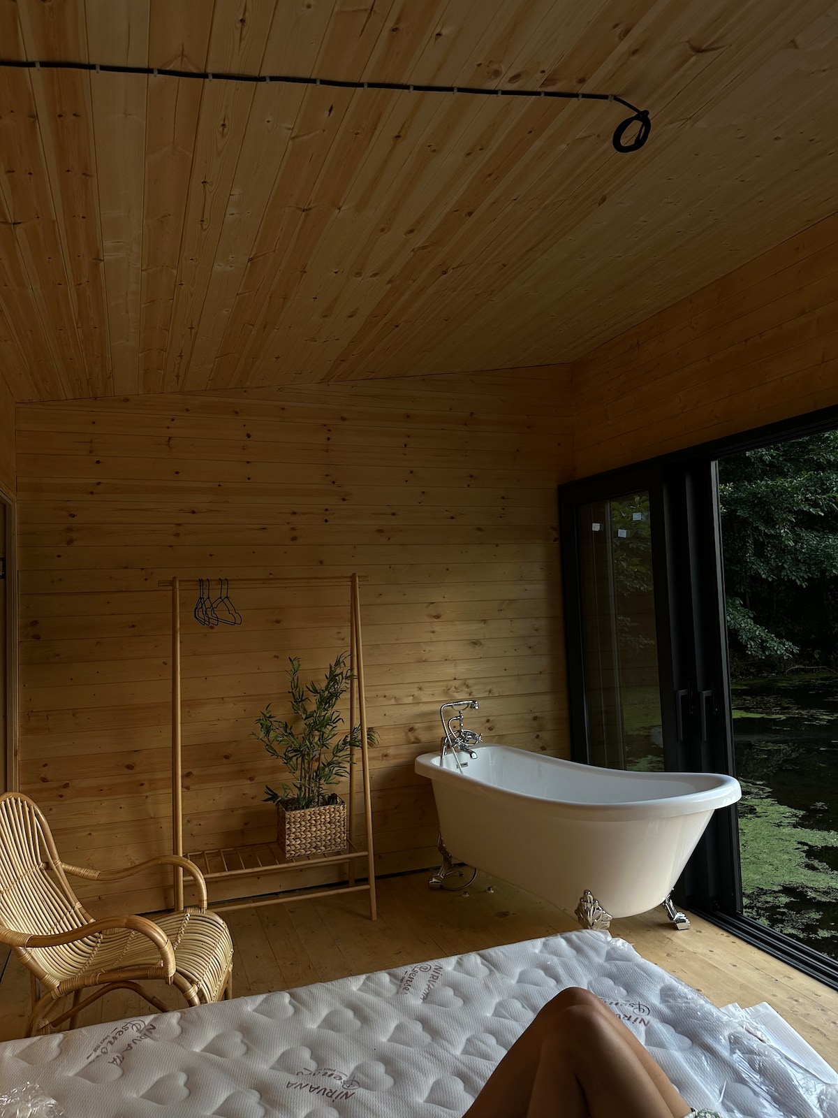 A serene bathroom features a freestanding bathtub next to large glass doors that offer views of lush greenery. The natural wood paneling and flooring create a warm atmosphere. A rattan chair is positioned nearby, complemented by a decorative plant in a woven basket.