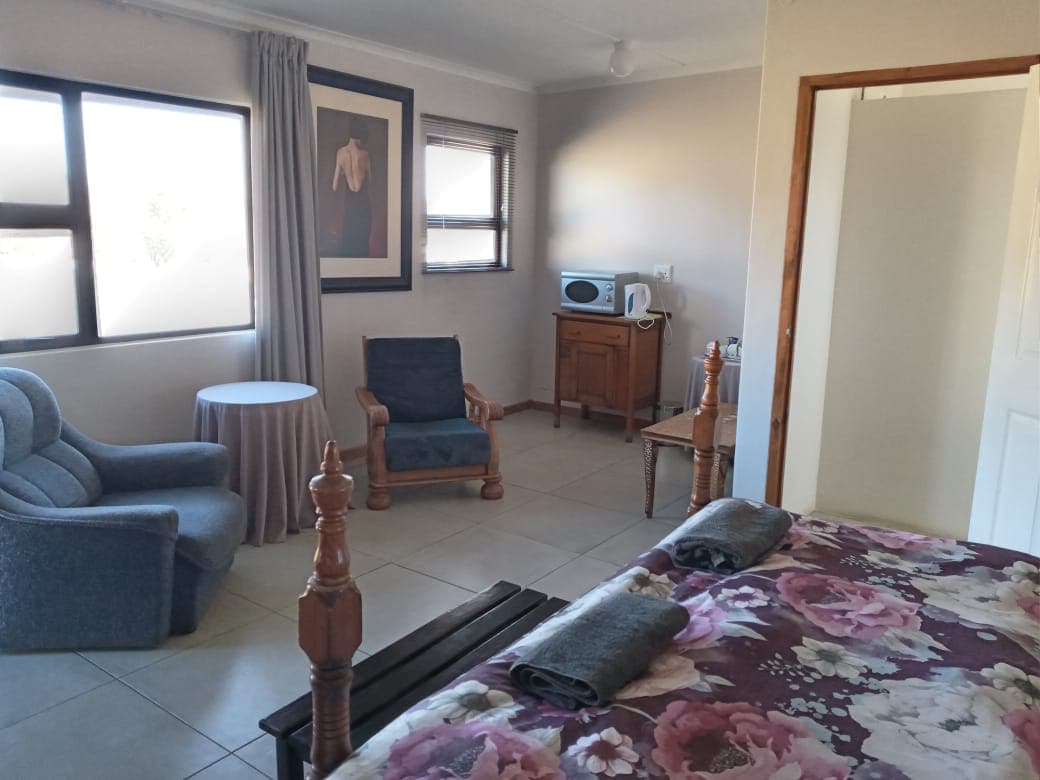 A comfortable sitting area is presented with two upholstered chairs and a small round table. A kitchenette is visible with a microwave and cabinet. Natural light enters through large windows, highlighting the floral-patterned bedding and wooden furnishings in the room.