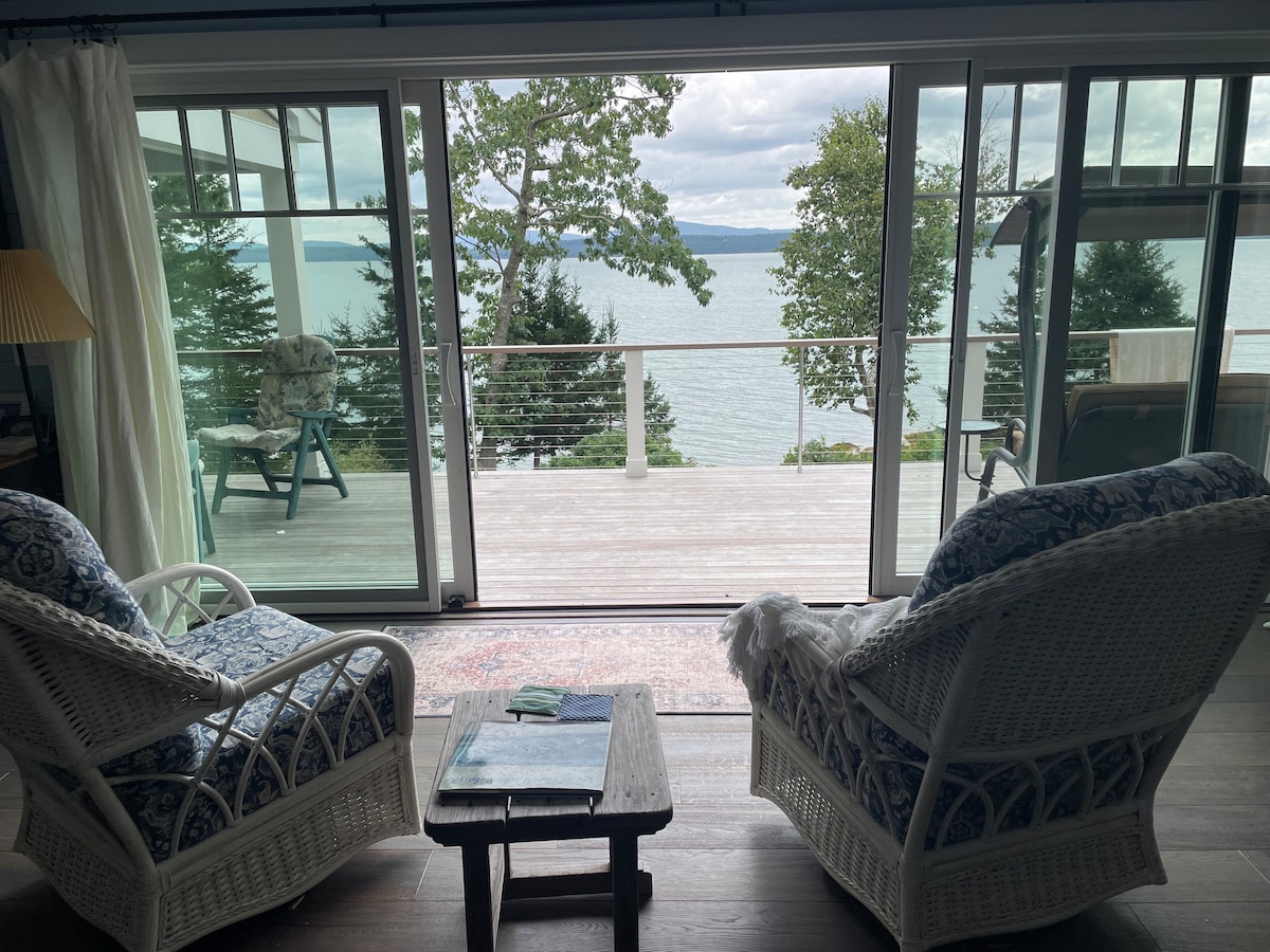 Two separate wicker chairs are positioned across from each other, facing large sliding glass doors. The doors open to a deck overlooking the water, framed by trees. A small table is situated between the chairs, with a light blue item resting on it.