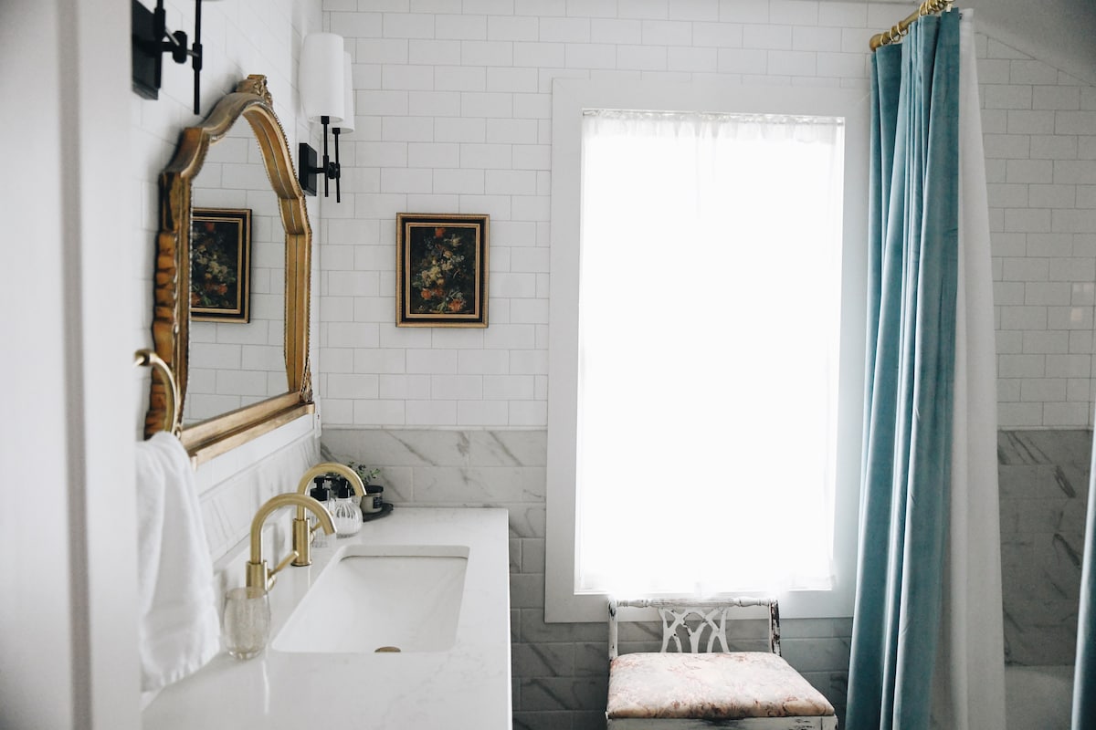 A clean and bright bathroom features a spacious sink area with brass fixtures. Natural light filters through a large window, accentuated by sheer white curtains. A vintage chair with a patterned cushion is positioned nearby, while a calming blue shower curtain adds a touch of color.