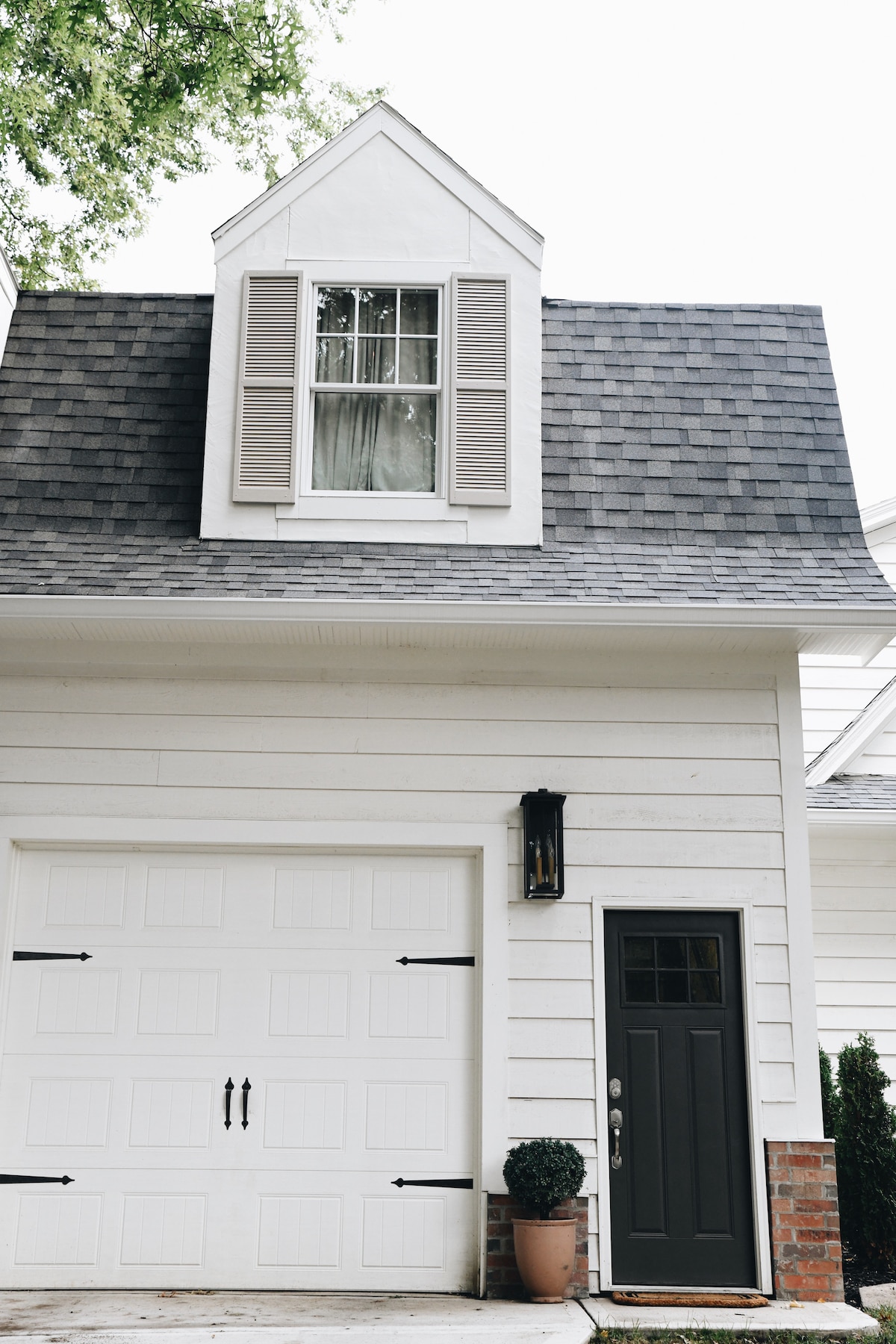 The exterior of a charming carriage house is displayed, featuring a pitched roof with a decorative dormer. Shuttered windows complement the light-colored siding. A garage door and a dark front door add contrast, while potted greenery enhances the welcoming entrance.