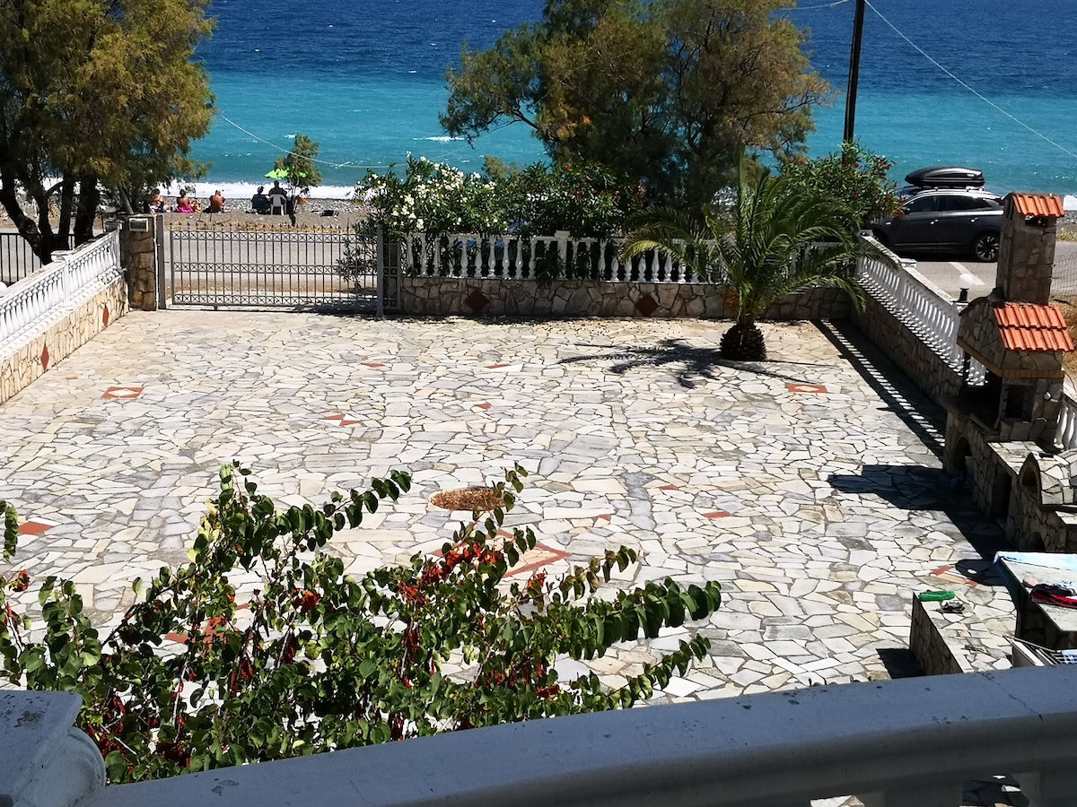 A spacious outdoor area is presented, featuring a stone pavement and surrounded by trees. A glimpse of the beach is visible in the background, along with some vehicles parked nearby. The property is bordered by a decorative fence, enhancing the overall appeal.