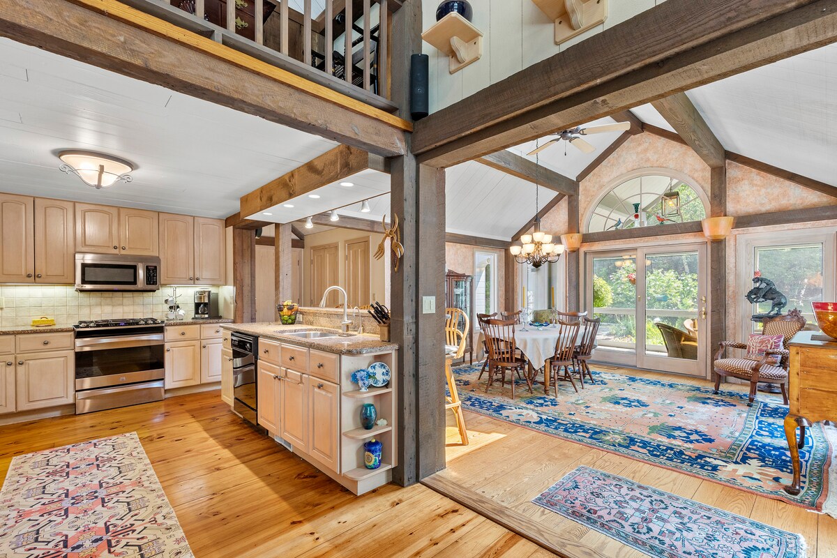 An open kitchen area features wooden cabinetry and modern appliances, with a large island for food preparation. Adjacent is a dining space with a round table surrounded by wooden chairs, complemented by large windows that frame views of the serene outdoor scenery.