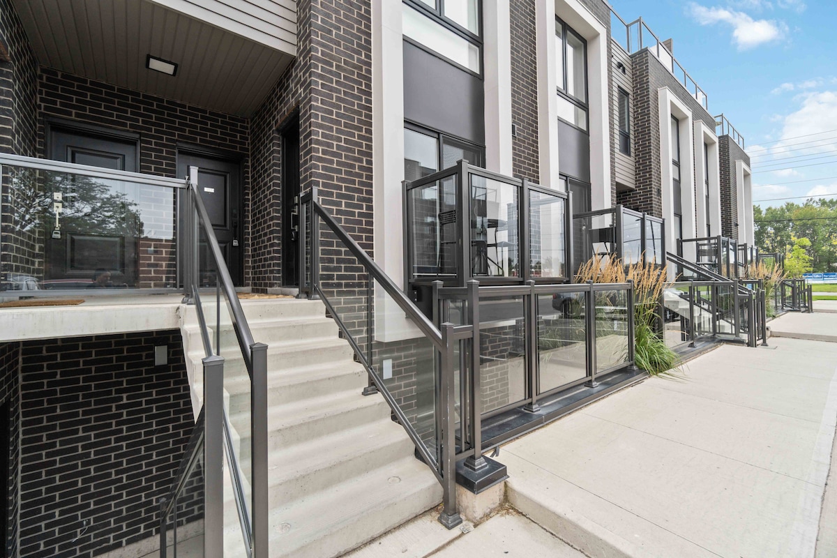 The exterior of a modern condo building showcases clear glass railings along the steps and walkways. A series of black and white facades create a sleek appearance. Green landscaping borders the entrance, contributing to the inviting aesthetics of the location.