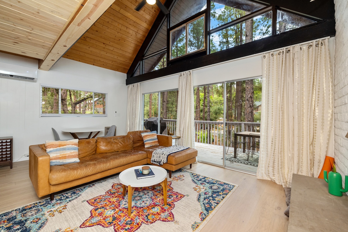 A spacious living area features a large leather sofa with decorative cushions, paired with a circular coffee table atop a colorful area rug. Floor-to-ceiling windows provide natural light and views of the surrounding trees, while sliding glass doors lead to a deck.
