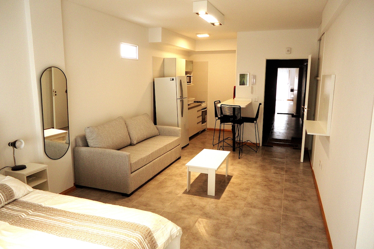 A modern living space features a light-colored sofa facing a small coffee table. A dining area with high stools is positioned next to a kitchen with essential appliances. Natural light enters through a nearby window, enhancing the room's spacious feel.