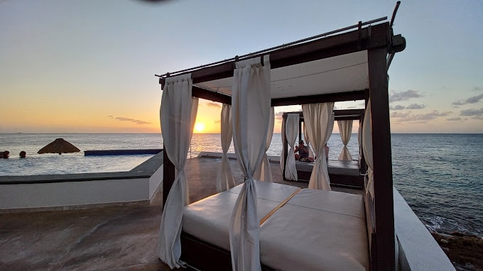 A serene poolside area is portrayed with comfortable cabanas draped in elegant white curtains. The setting sun casts a warm glow over the horizon, enhancing the tranquil atmosphere. The calm ocean is visible, creating a peaceful backdrop for relaxation.