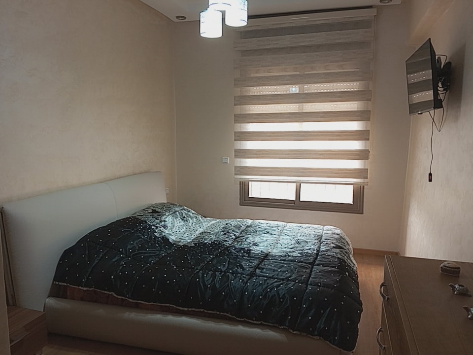 A comfortable bedroom features a spacious bed covered with a dark quilt patterned with small dots. Natural light filters through a horizontal striped window covering. A wall-mounted television is positioned opposite the bed, and minimalist wooden furniture complements the room's clean design.
