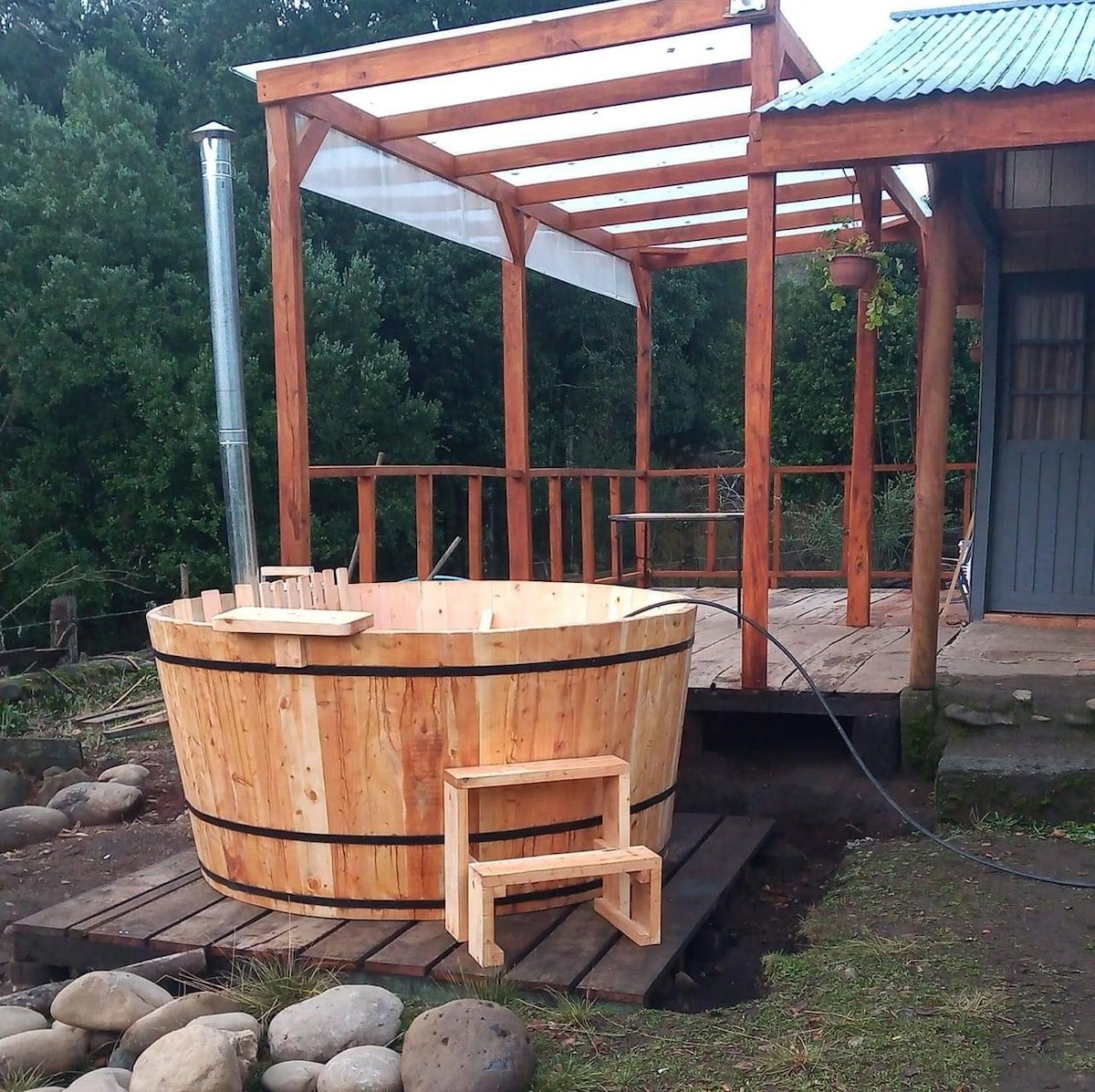 An outdoor wooden hot tub is situated on a deck made of natural wood. A small steps is positioned next to the tub. A partial roof covers the area, surrounded by lush greenery, providing a serene setting for relaxation.