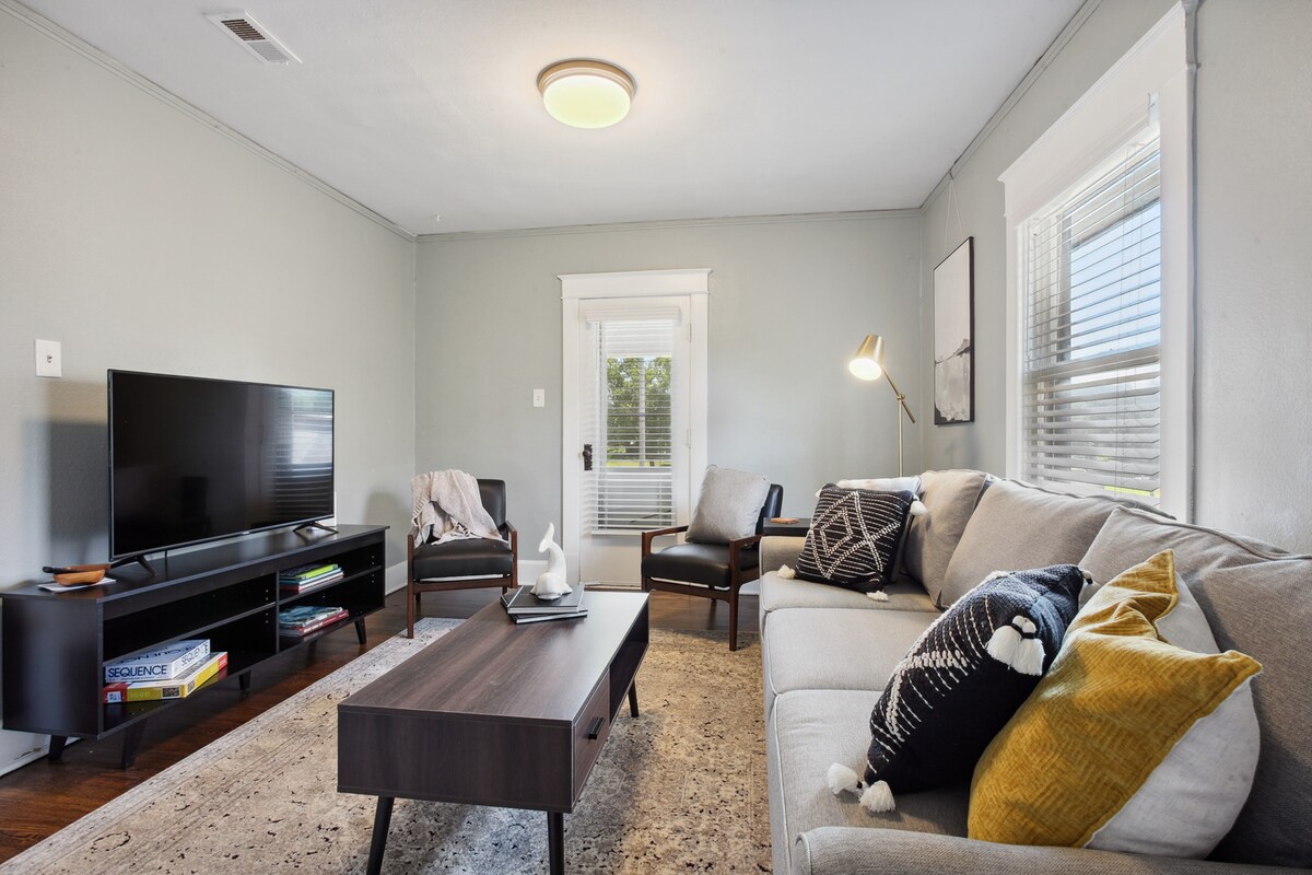 The living room features a comfortable sofa adorned with cushions, paired with an armchair. A smart TV is mounted on the wall across a dark coffee table, with a light rug underfoot. Natural light filters through the window, enhancing the welcoming space.