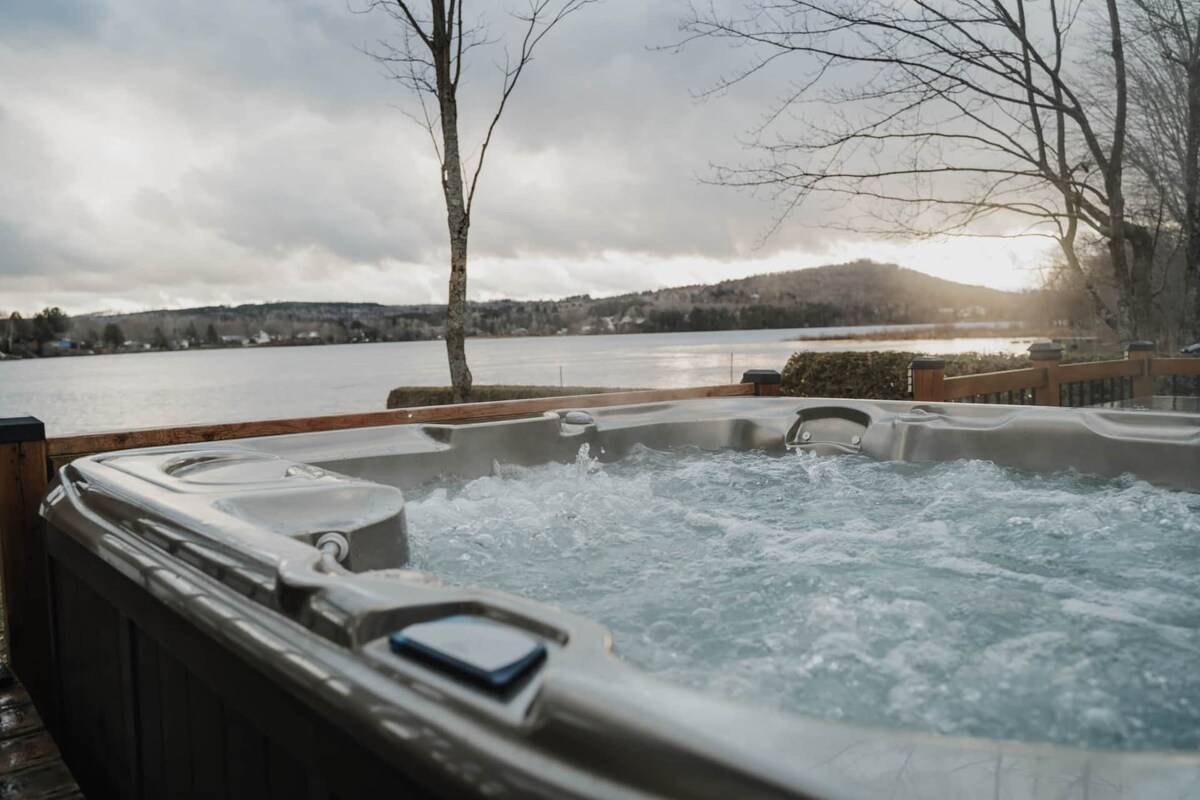 A hot tub filled with bubbling water is situated on a spacious deck overlooking a calm lake. Trees line the perimeter, and distant hills can be seen in the background under a cloudy sky, suggesting a peaceful outdoor retreat.
