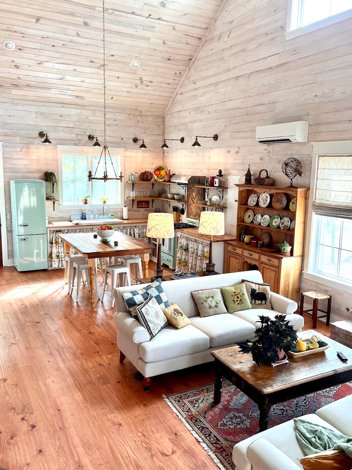 A spacious living area features a light wood ceiling with tall windows allowing natural light. A comfortable white sofa with decorative pillows faces a wooden coffee table. An inviting kitchen area with a dining table and vintage green refrigerator is seen in the background.