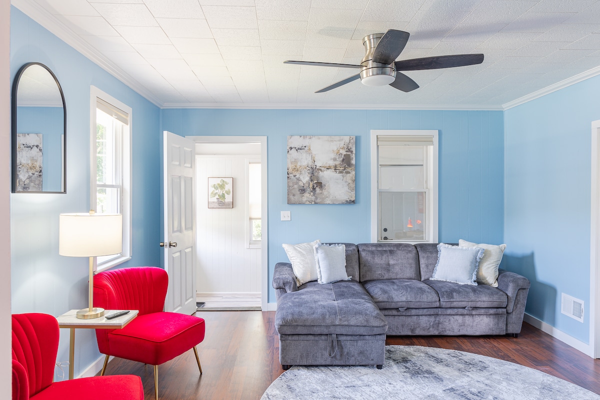 A bright living space is presented with a gray sectional couch and two red accent chairs. Natural light fills the room through a nearby window. A modern ceiling fan provides a cooling feature above, and a small table beside a chair holds a lamp, enhancing the inviting atmosphere.