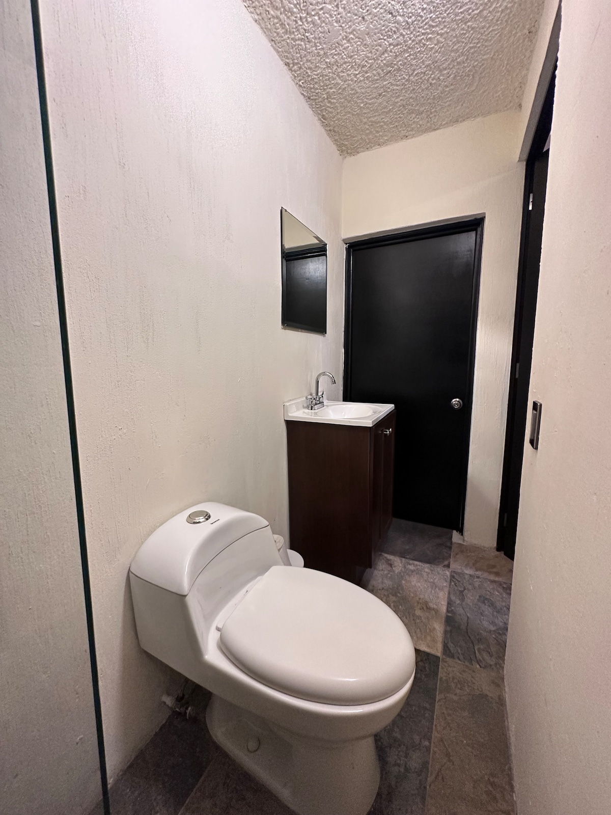 A clean and functional bathroom area is featured, showcasing a toilet and a small vanity with a sink. A mirror is mounted above the vanity. The flooring consists of large tiles, and the walls are lightly textured in a neutral tone.