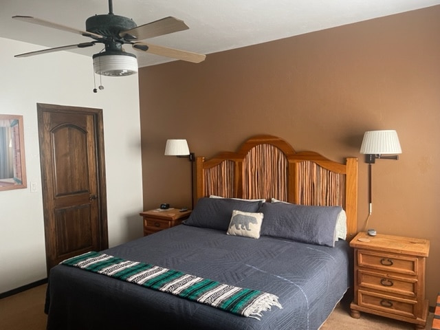 A spacious bedroom features a large wooden bed frame with a dark quilt and decorative throw blanket. Two bedside tables with lamps are positioned on either side. A ceiling fan is above, and a wall with a warm brown hue complements the natural wood elements.