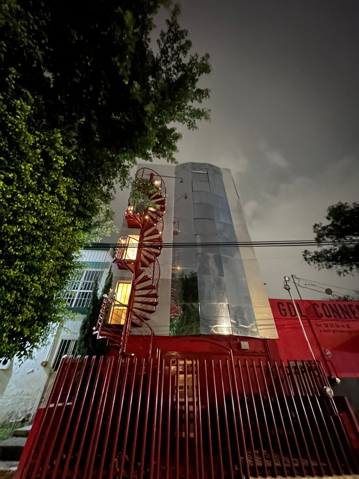 The exterior of a modern multi-story building is shown at night, featuring a metal facade that reflects nearby light. A prominent spiral staircase with greenery winds along the side, leading to upper levels. The entrance is framed by lush trees, enhancing the urban landscape.