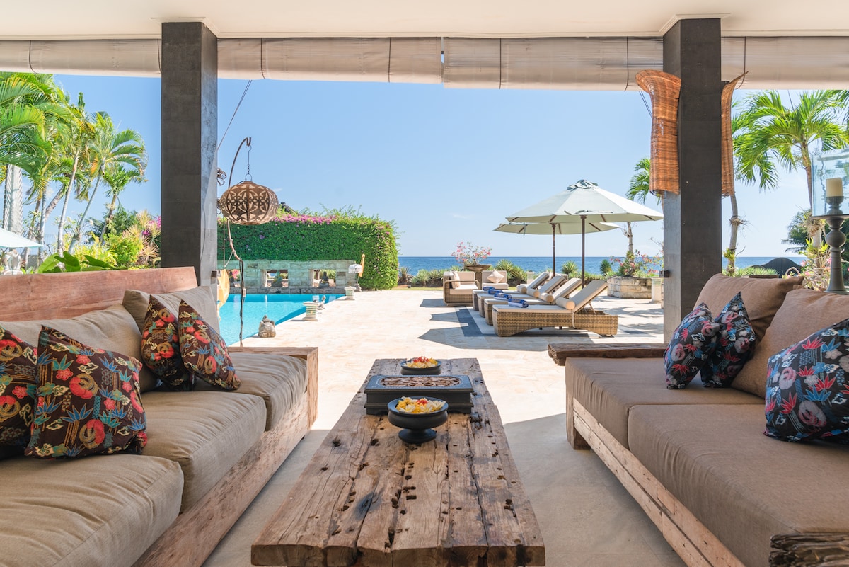A spacious outdoor lounge area features comfortable seating with colorful cushions and a rustic wooden table. Beyond, an infinity pool is visible alongside sun loungers and umbrellas, framed by lush tropical plants and the serene sea in the background.