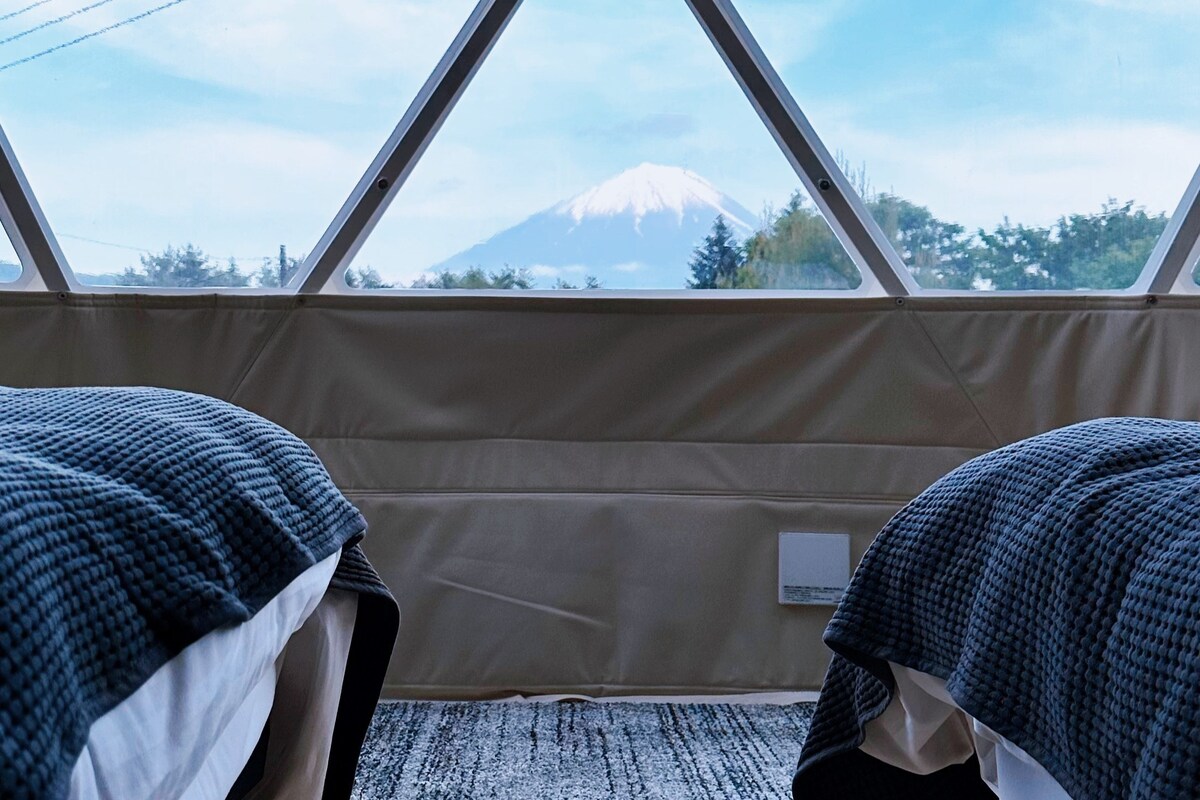 Two spacious beds are positioned side by side, with a notable view of Mount Fuji visible through large windows. The bedding features a textured pattern, adding to the inviting atmosphere. Natural light floods the room, enhancing the connection to the outside landscape.