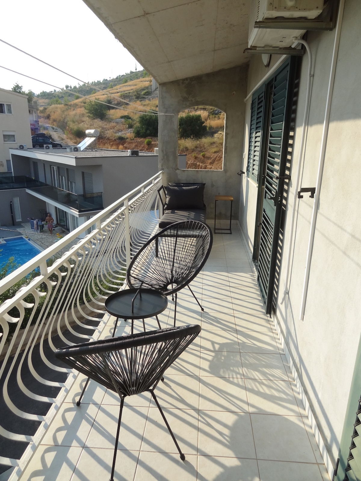 A spacious balcony features black Acapulco chairs and a small round table. The railing offers a view of the surrounding hillside. Sunlight casts gentle shadows on the tiled floor, enhancing the inviting yet relaxed outdoor space.