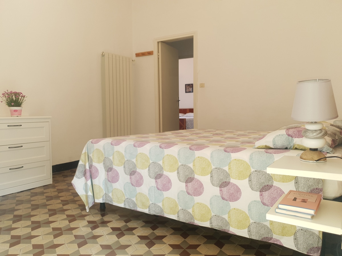 A well-lit bedroom features a spacious bed adorned with a patterned duvet. A white nightstand holds a lamp and a few books. A dresser with flowers brings a touch of color to the room. The floor is designed with a distinctive tile pattern.