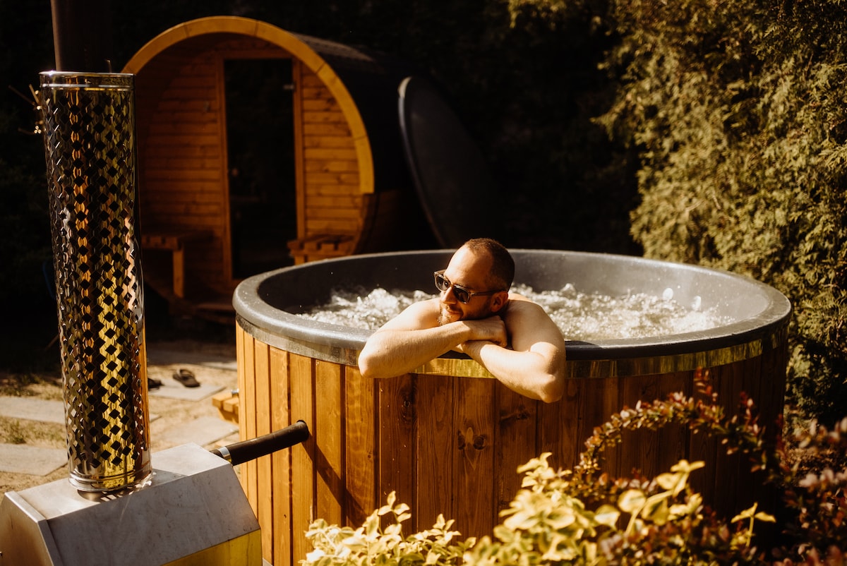 A wooden hot tub is surrounded by greenery, featuring bubbling water. Beside it, a wooden sauna structure is visible, complementing the serene outdoor setting. The scene conveys an inviting atmosphere for relaxation.