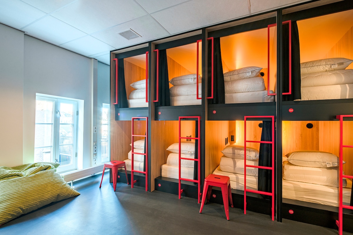 A spacious communal sleeping area features individual sleeping pods, each equipped with luxurious bedding and privacy curtains. Bright orange lighting illuminates the interior, while modern red accents highlight the structural design. A cozy bean bag chair is positioned near a window, providing comfortable seating.