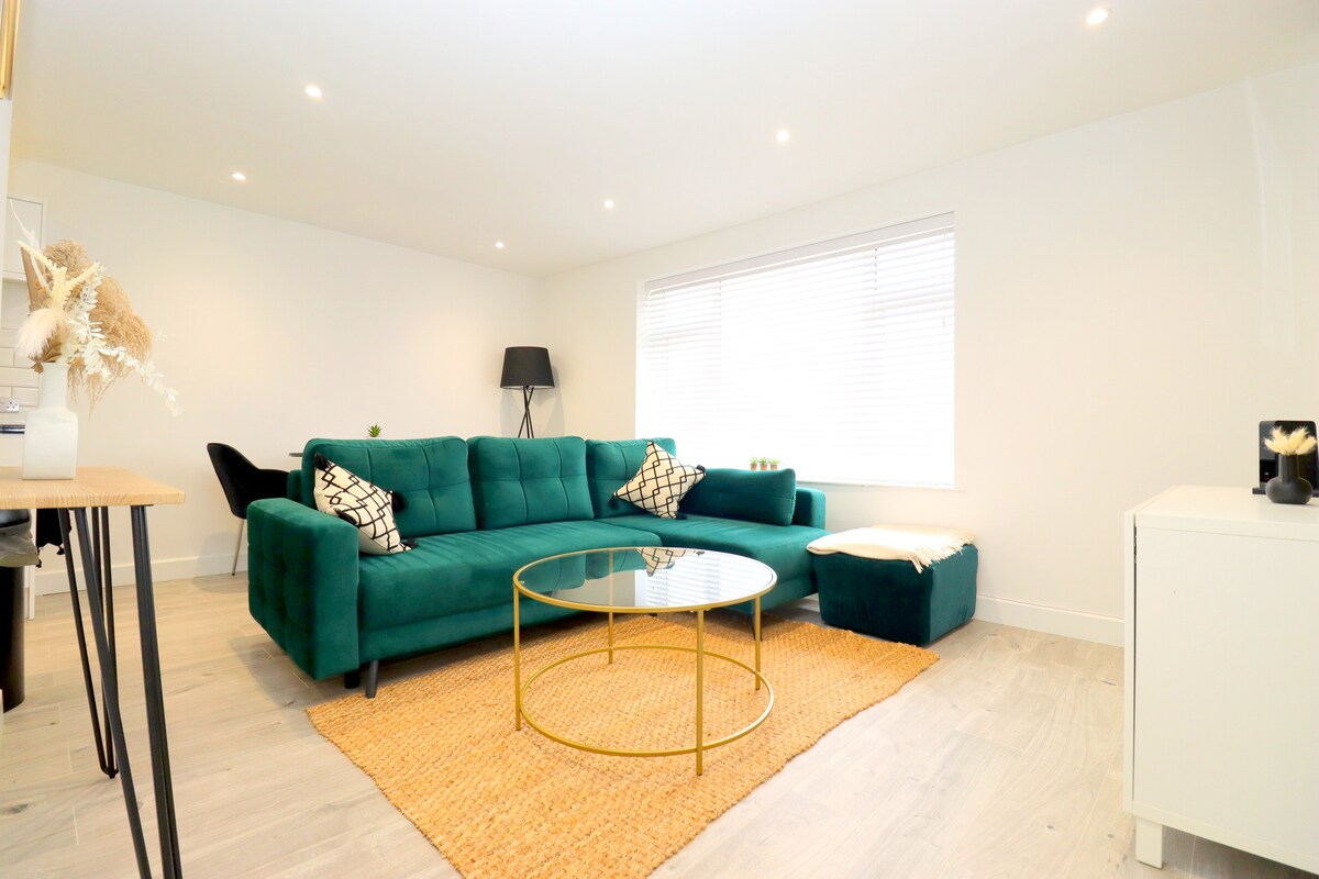 A modern living space features a plush green sofa paired with a round glass coffee table. Natural light fills the room, illuminating the neutral-colored walls and light wood flooring. A cozy throw blanket rests on a matching ottoman, while a standing lamp adds elegance.