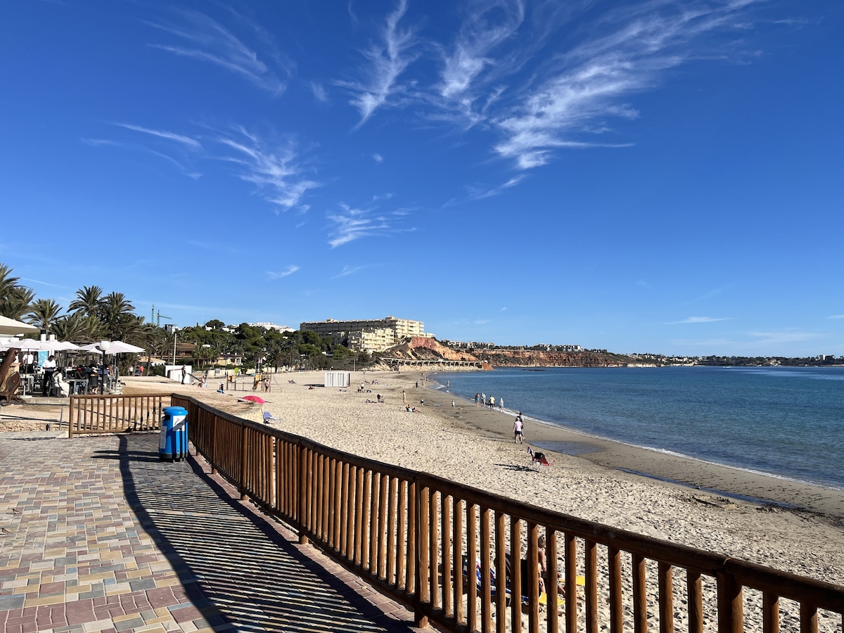The image shows a beach walkway lined with wooden railings, leading towards a sandy shoreline. In the distance, a coastal area with buildings and greenery is visible under a clear blue sky, while gentle waves meet the shore.