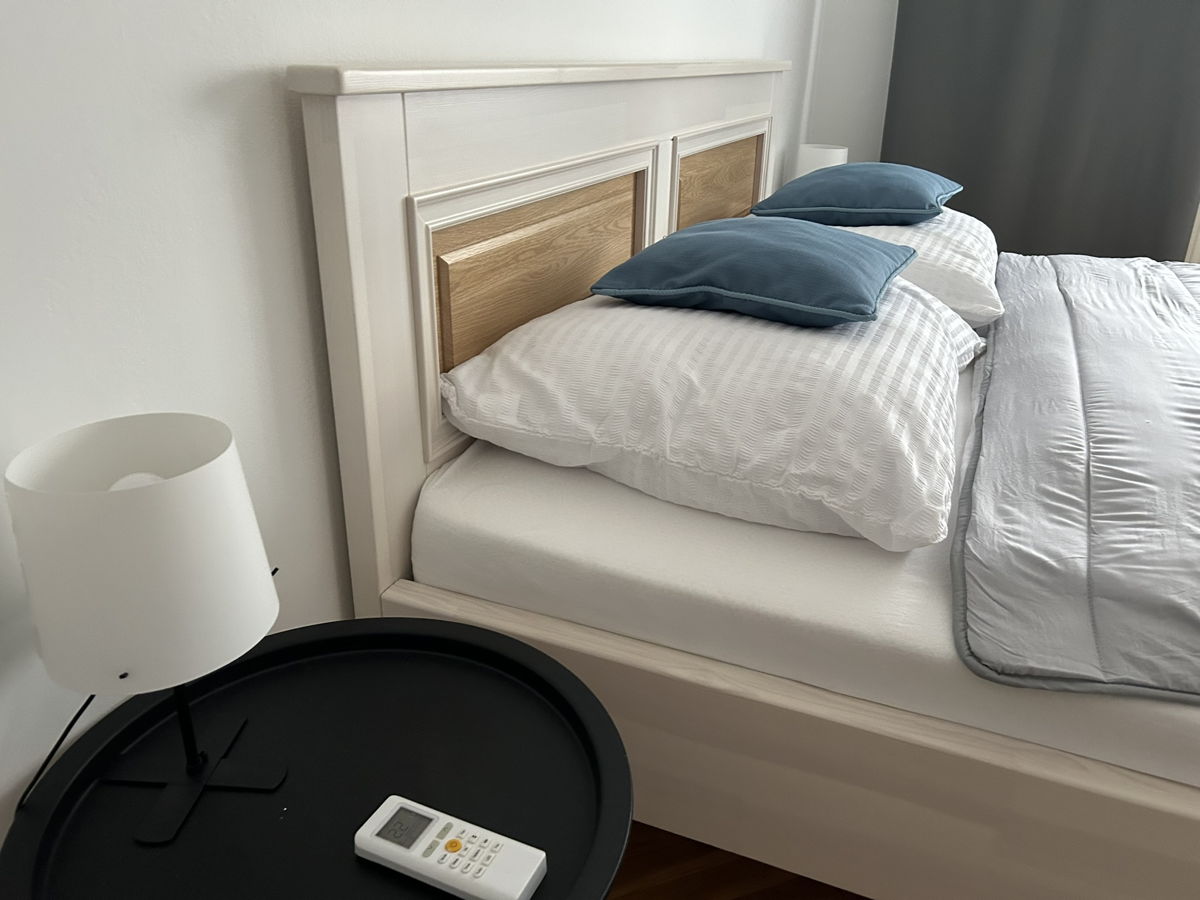 A spacious bed is presented, featuring a white wooden headboard and two large pillows in soft blue hues. A bedside table with a circular shape holds a remote control, while a minimalist lamp complements the decor. Natural light softly illuminates the area.