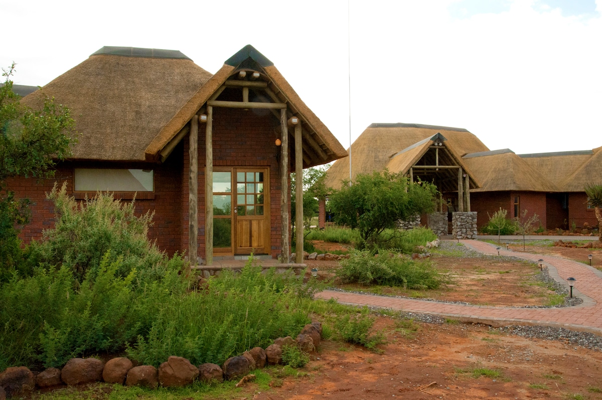 Chalets are set amid natural greenery, with thatched roofs and brick walls blending harmoniously into the landscape. A welcoming wooden door is visible, framed by surrounding plants. A stone pathway connects the structures, enhancing the sense of privacy and tranquility.