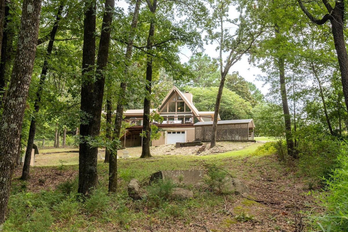 A spacious A-frame home is nestled among tall trees, emphasizing its natural setting. The structure features large windows that stretch from the roof to the ground, providing a connection to the surrounding greenery. A gentle slope leads up to the home's entrance.