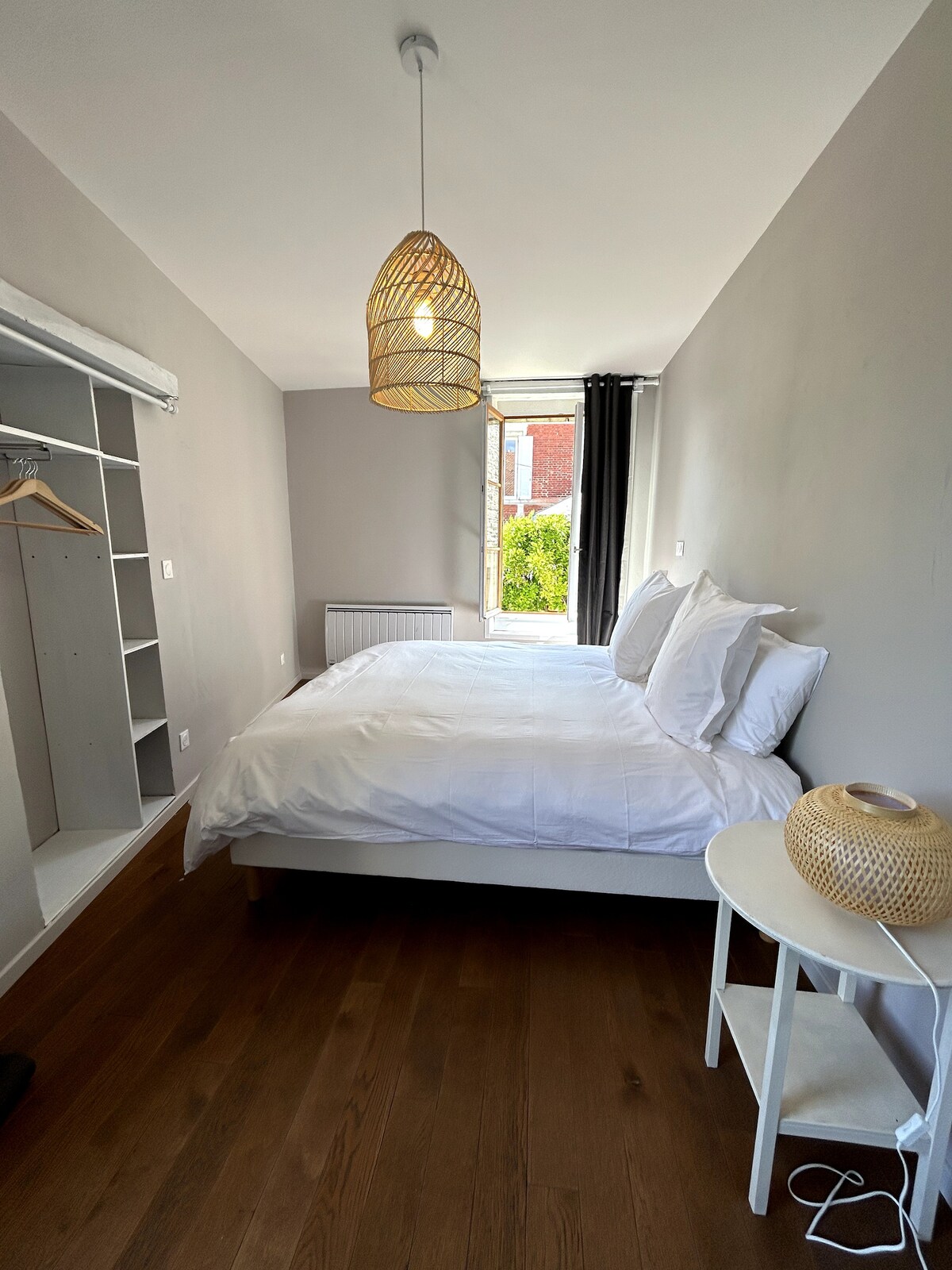 A bright bedroom features a spacious bed with crisp white linens. A woven pendant light hangs above, and a small round table sits beside the bed. A closet with hanging space is visible, and natural light enters through a window dressed with dark curtains.