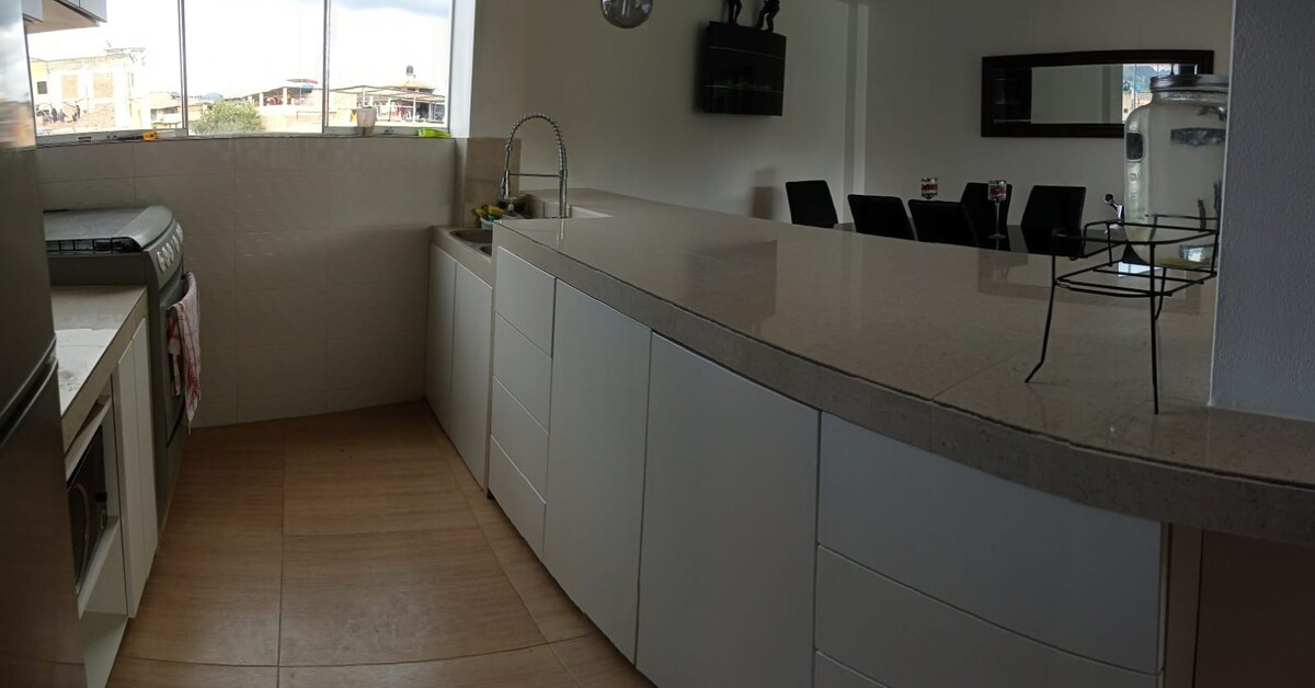 A modern kitchen is displayed, featuring sleek white cabinetry and a spacious countertop. A sink is positioned near a large window, allowing natural light to fill the area. A dining set is visible in the background, suggesting a practical layout for meal preparation and dining.