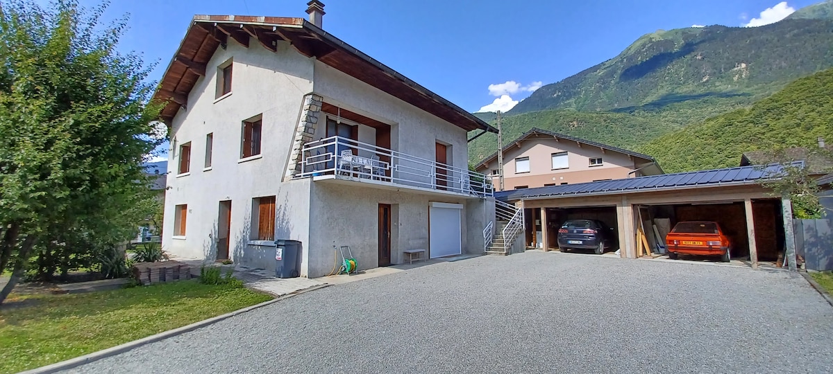 The exterior of the apartment features a two-story house with a balcony and a spacious parking area in front. Two vehicles are visible parked in the covered area on the right. Mountain views are visible in the background, contributing to a serene environment.