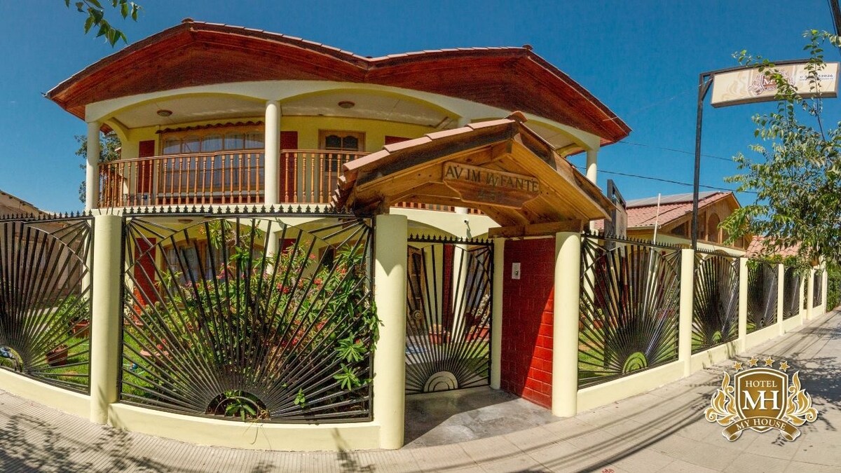 The exterior of the hotel features a distinctive entrance with a wooden roof and ornate metalwork. The vibrant facade is framed by lush greenery, creating a welcoming appearance. A tiered balcony is visible above the entrance, enhancing the building's architectural character.