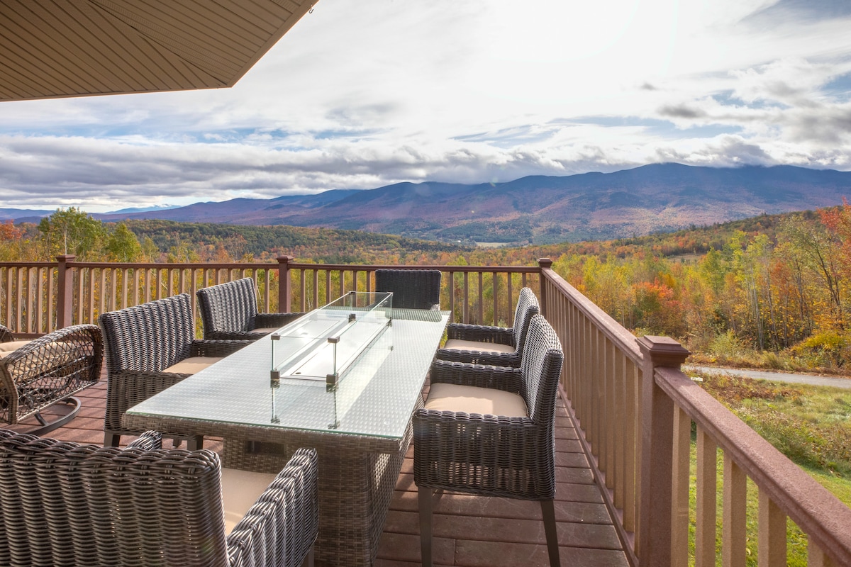 An outdoor dining area features a glass table surrounded by wicker chairs, situated on a wooden deck. Expansive views of rolling hills and mountains are visible in the background, showcasing a vibrant display of autumn foliage under a partly cloudy sky.