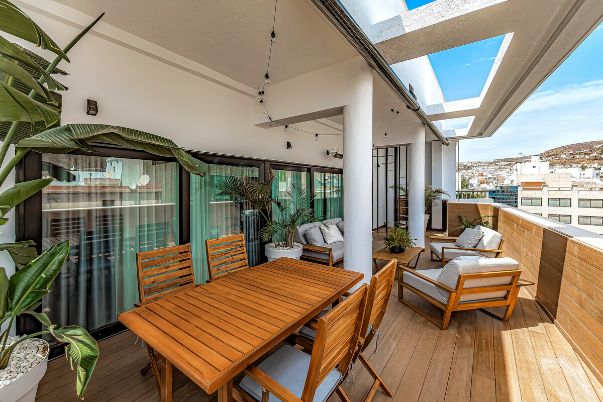 An outdoor terrace is furnished with a wooden dining table surrounded by matching chairs. Relaxing lounge areas are positioned with plush seating. Potted plants add greenery, while large windows provide expansive city views and ample natural light.