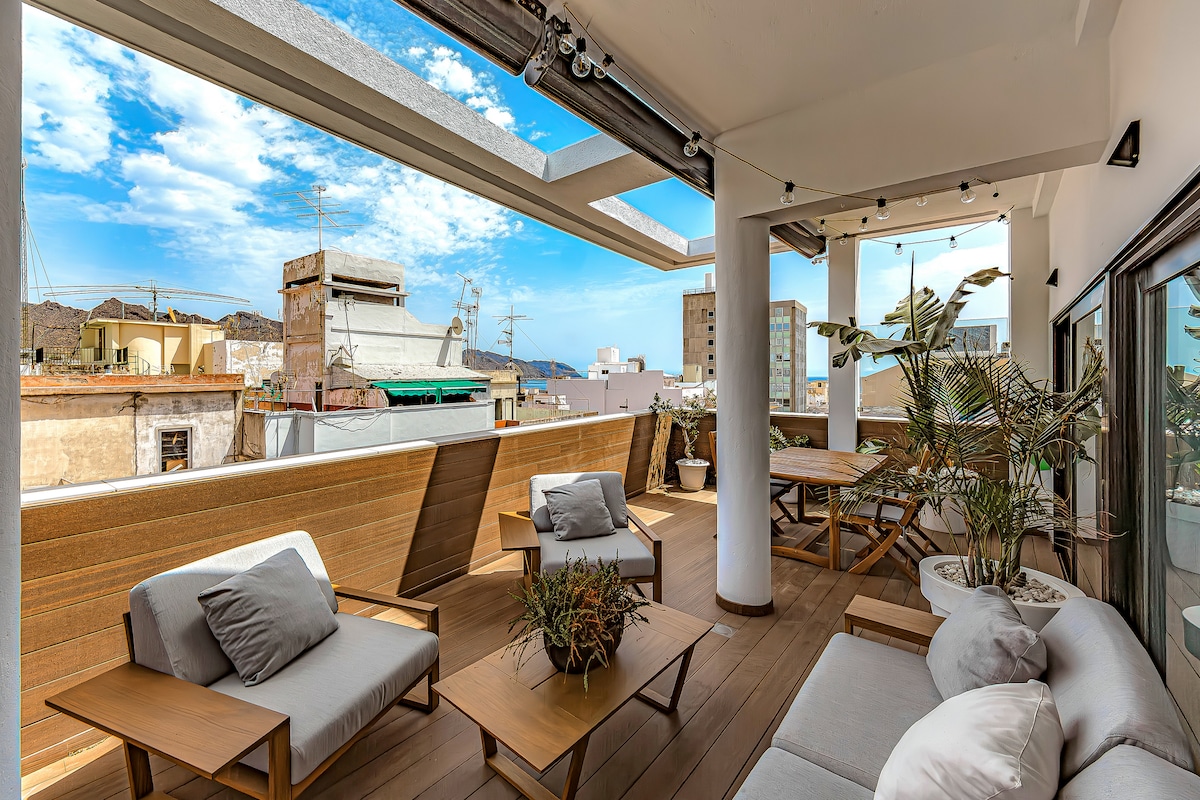 A spacious terrace is highlighted with comfortable seating arrangements, including gray sofas and wooden tables. Planters with greenery add a touch of nature. The sky above features a mix of clouds and bright blue, enhancing the vibrant outdoor setting.