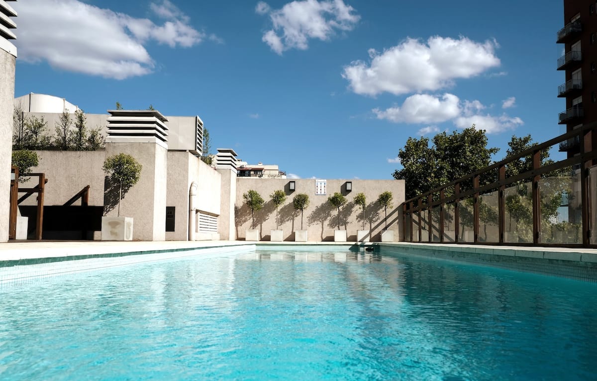 The rooftop pool reflects the clear blue sky, bordered by minimalistic landscaping and a wooden railing. Sunlight creates a tranquil atmosphere over the shimmering water, inviting relaxation and leisure.